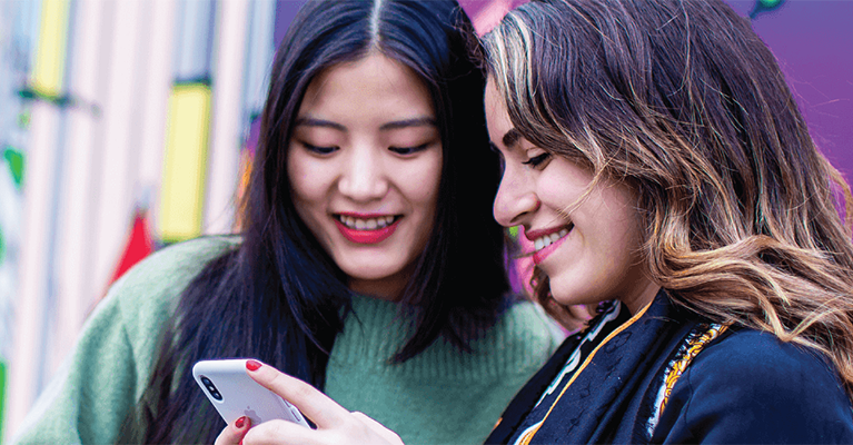 Two female students smile at what is on the screen of a phone.